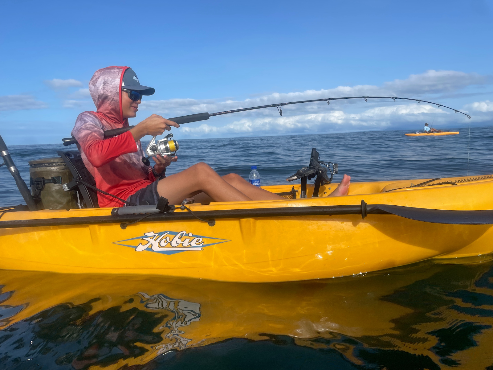 A wide shot of two yellow fishing kayaks floating on calm turquoise ocean water under a partly cloudy sky, with a lush green, mountainous tropical coastline in the background. The scene is bright and conveys a sense of peaceful morning adventure on the Pacific coast.