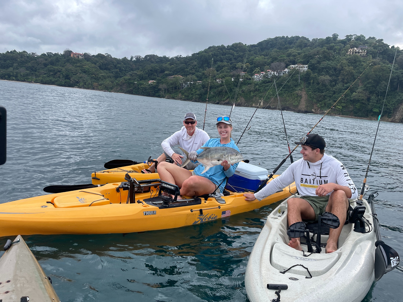 Three friends sitting on colorful Hobie fishing kayaks in the ocean, smiling and holding up a caught fish. The kayaks are equipped with gear, and the background features a lush, hilly coastline under a slightly overcast sky, highlighting a fun group fishing experience.