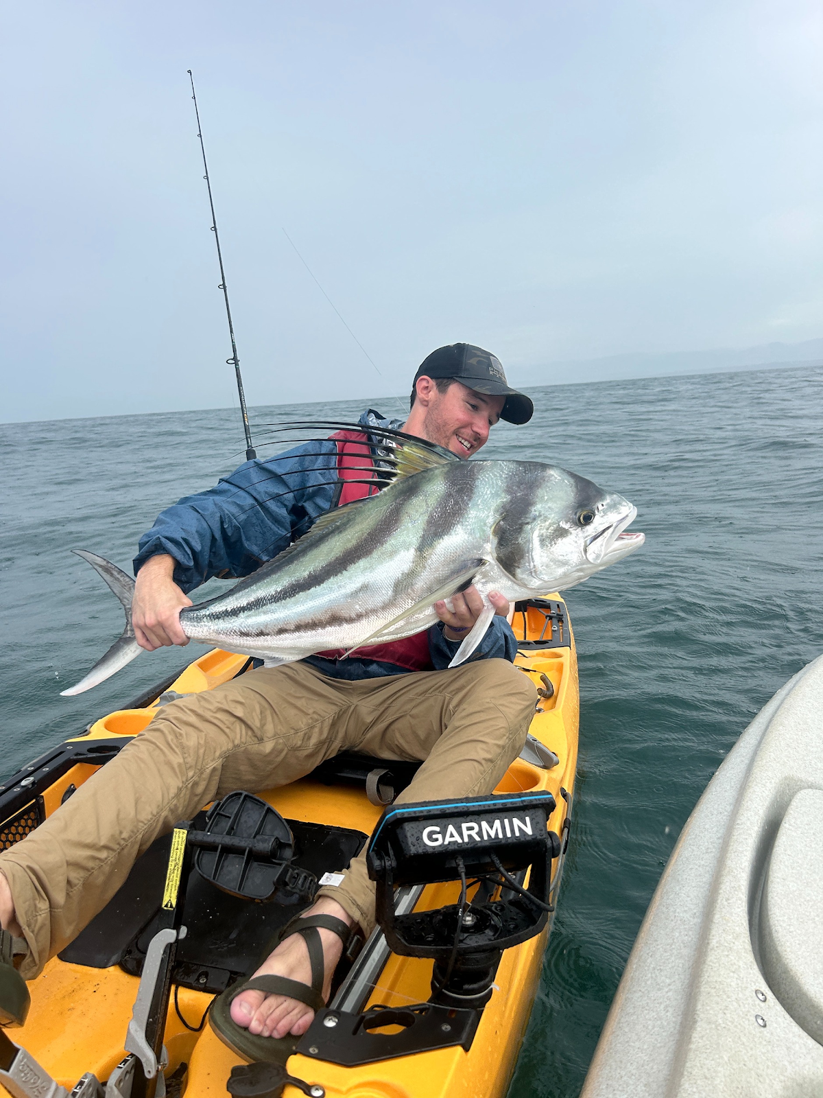 Ryan sitting in a yellow Hobie kayak holding a large fish, smiling, with the ocean and coastline behind him.